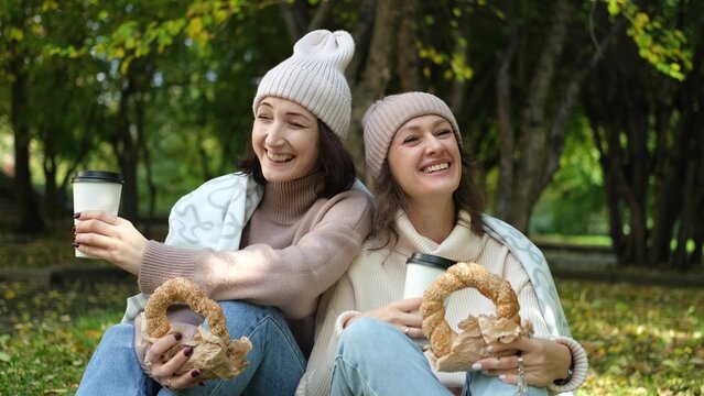 Two cute women in warm clothes sit hugging in an autumn park. Girlfriends enjoying beautiful weather, drinking tea or coffee, eating fresh bagels from the bakery