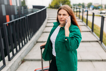 Fototapeta premium Young unhappy redhead businesswoman with red suitcase making phone call over mobile phone in the city. Portrait of a furious woman yelling at smartphone. Negative people emotion.