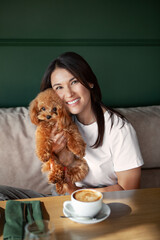Beautiful smiling Woman holds small dog in hands sits at table in restaurant with cup of coffee. Dark hair female in cafe with pet miniature poodle on dark background