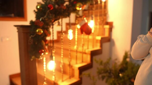 A woman gives her friend a Christmas gift, that she made with her own hands, against the background of a staircase decorated for Christmas in the house