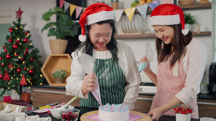 Happy asian elderly mother and young daughter helping squeezes cream onto a cake together. Mother and daughter making cake for Christmas day. Christmas cake decorated. Festive holiday