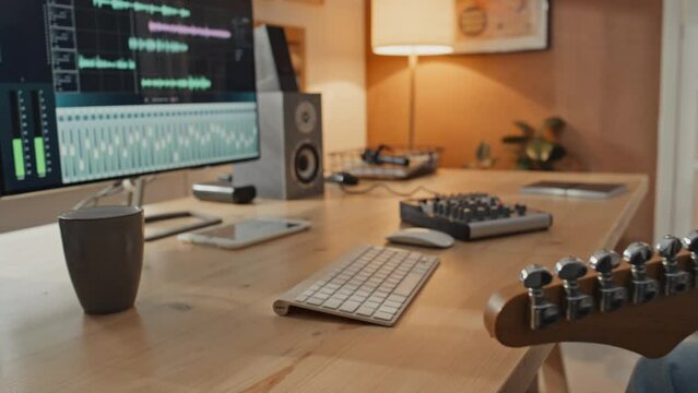 Close-up shot of young male musician sitting at table at home with computer, turning on recording software and mixer, putting on headphones and starting to compose new song on electric guitar