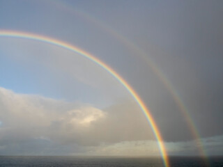 Colorful and dramatic double rainbow forms a full arch over the wake of a departing cruise ship at sea