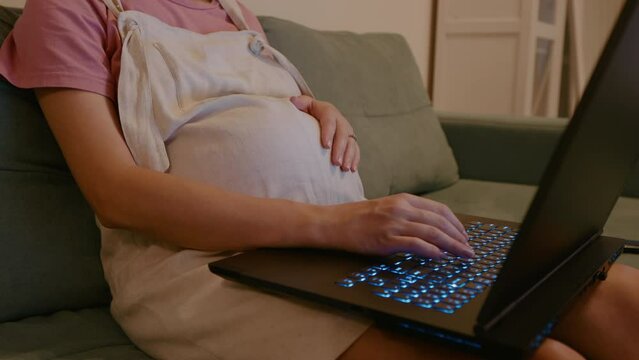 Side view medium shot of hands of pregnant woman who is using the keyboard laptop working while on maternity leave. Pregnant woman freelancing on a laptop close-up.