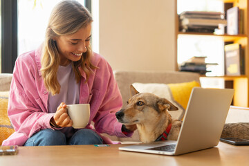 Smiling beautiful woman sitting on sofa, using laptop, holding cup, stroking pet, dog, at home