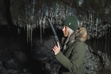 A girl in a hat and parka with a pistol with a silencer in winter near the ruins.