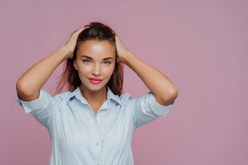 Chic woman with hands in hair, wearing a light blue shirt, with a confident look on a pink background