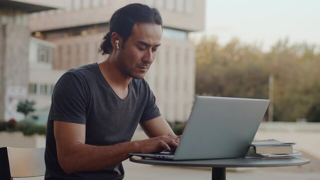 Young asian man in wireless headphones close up sitting at the outdoor cafe with a focused face and working on a laptop with a building in a backdrop. Concept of modern remote work with a flexible