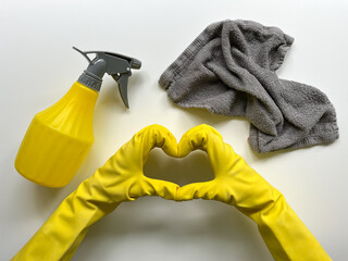 Yellow spray, grey cleaning cloth, and hands in rubber gloves make a heart on a white background. 