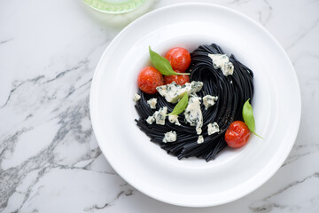 White plate with squid-ink spaghetti, blue cheese and tomatoes, horizontal shot on a white marble background, high angle view