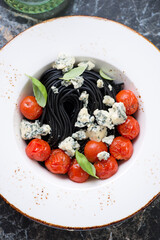 Beige plate with black spaghetti, blue cheese, roasted cherry tomatoes and basil, top view on a black marble surface, vertical shot, closeup