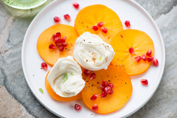 Persimmon slices with torn mozzarella ball and pomegranate seeds on a white plate, high angle view, middle close-up