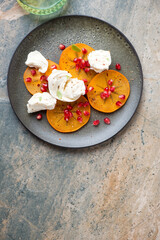 Plate with fresh sliced persimmon, torn mozzarella and pomegranate, top view on a beige and grey granite background, vertical shot with space