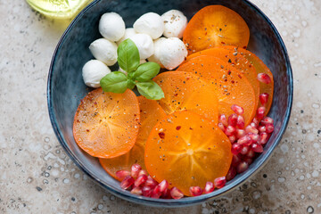 Blue bowl with sliced persimmon, mozzarella balls and pomegranate seeds, middle close-up on a beige granite background, horizontal shot