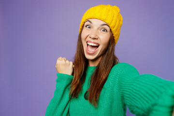 Close up young woman she wear green sweater yellow hat casual clothes doing selfie shot pov on mobile cell phone do winner gesture isolated on plain pastel light purple background. Lifestyle concept.