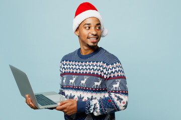Side view young IT man wear knitted sweater Santa hat posing hold use work on laptop pc computer look aside isolated on plain blue background Happy New Year 2024 celebration Christmas holiday concept
