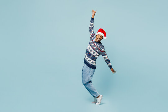 Full body young man wear knitted sweater Santa hat posing stand on toes with outstretched hands lean back isolated on plain blue background. Happy New Year 2024 celebration Christmas holiday concept.
