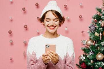 Merry young woman wear white sweater hat posing hold in hand use mobile cell phone typing message...