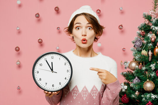 Merry surprised young woman wear white sweater hat posing hold in hand point index finger on clock isolated on plain pastel pink background studio Happy New Year celebration Christmas holiday concept