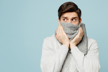 Young ill sick man wear gray sweater cover mouth with scarf look aside on area isolated on plain blue background studio portrait Healthy lifestyle disease virus treatment cold season recovery concept