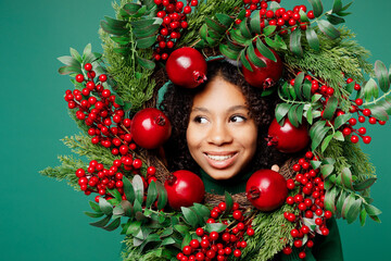 Close up smiling cheerful merry little kid teen girl posing hold looking aside through Christmas handmade wreath isolated on plain green background studio. Happy New Year celebration holiday concept.