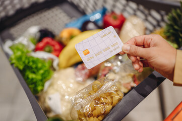 Close up cropped customer woman hold credit bank card near trolley cart, buying goods shopping at supermaket store grocery shop choosing products inside hypermarket Purchasing food gastronomy concept