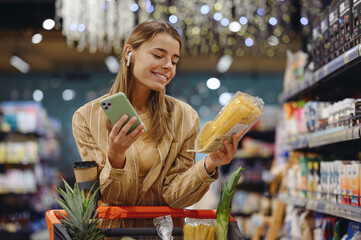 Young woman wears casual clothes earphones listen music use mobile cell phone hold pasta shopping at supermaket store grocery shop buy with cart choose products in hypermarket Purchasing food concept