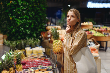 Young minded customer woman wear casual clothes hold pine-apple shopping with shopper bag at supermaket store grocery shop buy fruit choose products in hypermarket. Purchasing food gastronomy concept.