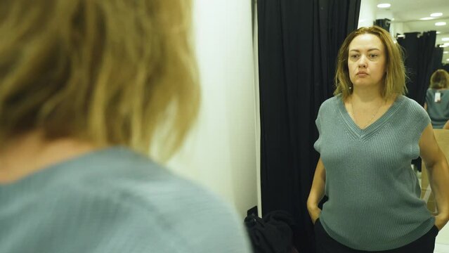 A woman tries on a jumper and looks in the mirror. Selecting and purchasing clothes in a store.