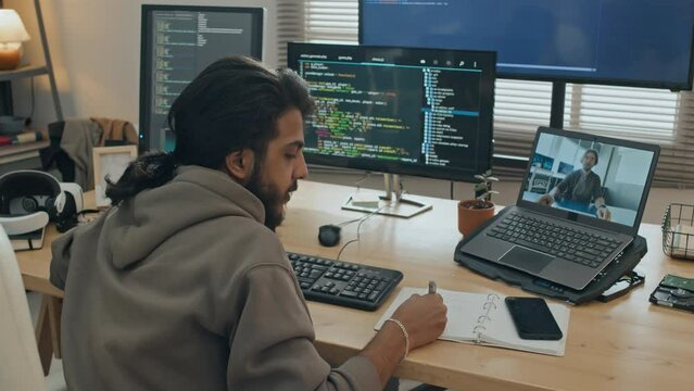 Medium rear shot of male software developer sitting at desk with several computer screens, talking to colleague on video call on laptop, discussing issue and arguing, while working at home
