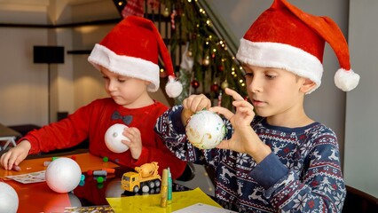 Portrait of two boy painting and decorating Christmas baubles with glitter. Winter holidays, family time together, kids with parents celebrating.