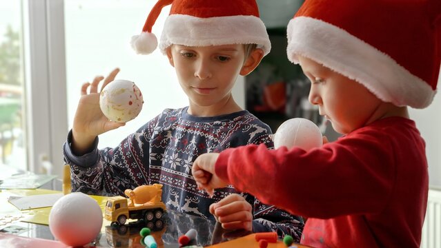 Portrait of two cheerful boy in Santa's hats being creative while making traditional Christmas decorations, garlands and baubles. Winter holidays, family time together, kids with parents celebrating.