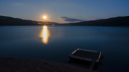 Under the glow of a full moon, a serene lake unveils its calm waters. A wooden pontoon stretches gracefully near the shore. Summer nights in the mountains. Izvorul Muntelui Lake, Romania. - obrazy, fototapety, plakaty