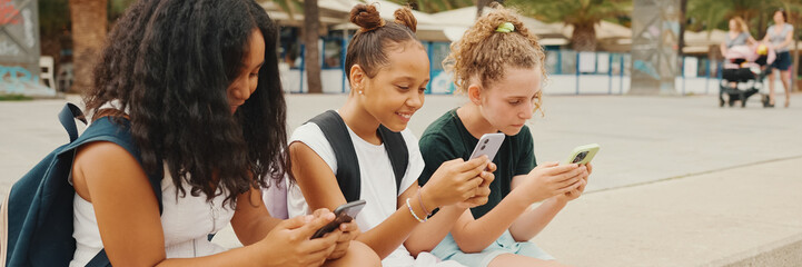 Three girls friends pre-teenage are sitting on the waterfront using mobile phone. Three teenagers playing games on a smartphone on the outdoors in urban cityscape background