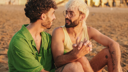 Romantic moments, homosexual couple enjoying communication with each other while sitting on the beach at sunrise, crossing their arms and looking at each other with love