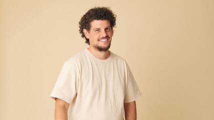 Fototapeta na wymiar Happy guy with curly hair dressed in beige t-shirt laughing looking at camera isolated on beige background in studio
