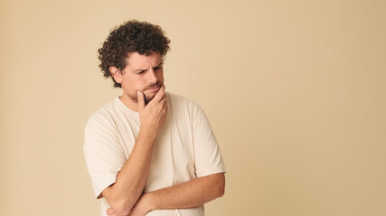 Thoughtful guy with curly hair dressed in beige t-shirt, with his hands folded unable to make up his mind in studio on beige background in the studio