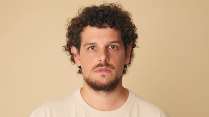 Close-up of an amazed guy with curly hair dressed in beige t-shirt looking at the camera with big eyes expressing wow while standing in studio on beige background in the studio - obrazy, fototapety, plakaty