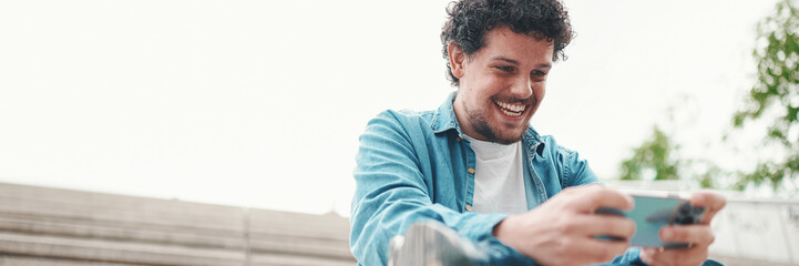 smiling young bearded man in denim shirt sitting on high steps and using mobile phone. Man playing on smartphone