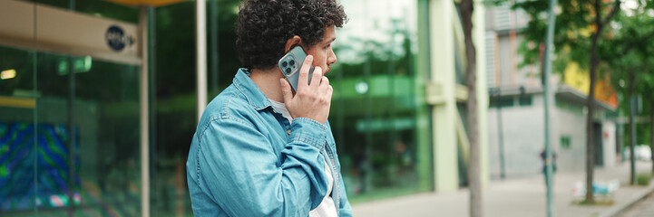 young bearded man in denim shirt stands at bus stop waiting for bus and talking on mobile phone on...