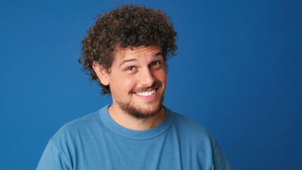 Close up, pensive guy with curly hair dressed in blue t-shirt looks at the camera having good idea...