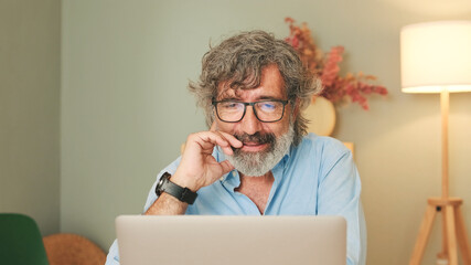 Happy senior man with gray hair wearing glasses, in living-room making online video call using laptop talking