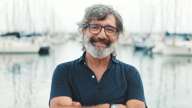 Happy male seigneur in glasses crossing his arms while standing in the port and looking at the camera on yachts background, camera is moving closer
