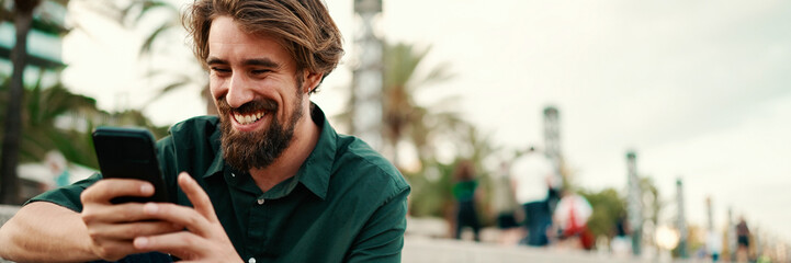 Fototapeta premium Close-up portrait of a man with a beard recording a voice message on the embankment background. Closeup of a young hipster male using a mobile phone