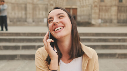 Close up, happy traveler girl talking on mobile phone while sitting on the steps of an old building in the historical part of an old European city