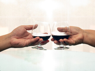 Close up two people holding and toast wine glass together near window light with white curtain
