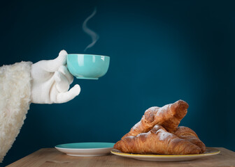 Santa Claus hand holding a cup of coffee on a turquoise background. There are delicious croissants with powdered sugar on the table.