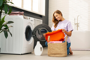 Beautiful smiling blonde woman, housekeeper holding fresh and clean  clothes sitting near washing machine  sitting at home. Laundry concept