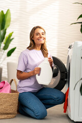 Beautiful smiling young woman holding bottle with laundry detergent sitting near washing machine...