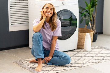 Young smiling confident woman sitting on carpet looking at camera in modern laundry room with...